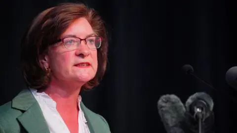 PA Media A woman with short dark hair, wearing glasses, a white blouse and green suit jacket. She is stood in front of microphones and looking nto the distance with a very serious expression.