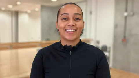 BBC Women's sport advocate Demi Brown stands facing the camera, smiling. She is wearing a zip-up, long-sleeve black sports top.
