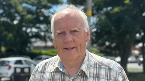 Alan Graham is wearing a checked shirt and is smiling at camera. He has white hair at the sides of his head and is balding on top. he is standing in front of a car park with some cars parked.