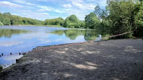 Derbyshire County Council Bank of large pond blocked off with hazard tape