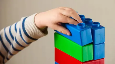 A child's hand holds blue plastic brick on a pile of other bricks.