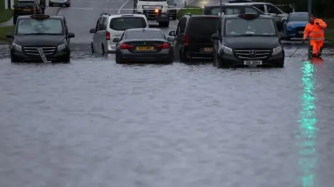 Reuters Seorang pekerja dewan mencoba membuka sumbatan saluran air setelah hujan lebat semalaman menyebabkan jalan terendam banjir, menyebabkan mobil-mobil terdampar di Manchester