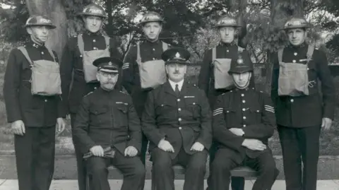A group of men in wartime police uniform, some sitting and others standing.
