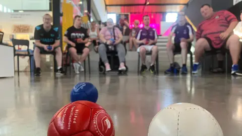 Jon Wright/BBC Six boccia players are seated in the background. in the foreground are a red, white and blue ball.