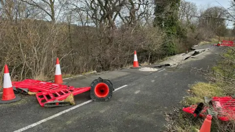 Cones and fallen barriers surround the section of road which is slipping into the River Wear.