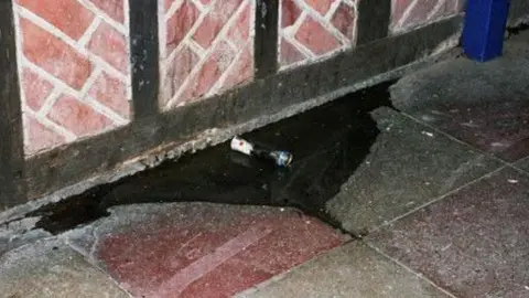 Sussex Police A cigarette butt on the floor of a concrete slabbed floor. The wall has red brick patterns on it. The cigarette butt is sat in a pool of liquid.