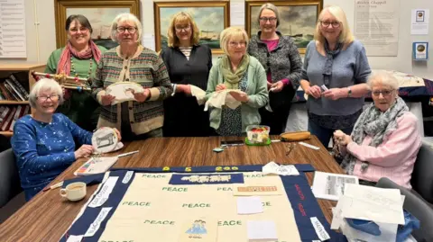 The photo shows eight women smiling at the camera. They are all holding pieces of fabric that they are embroidering. In front of them is a table, which has a large piece of fabric laid out on it. There are a collection of gold photo frames behind them. 