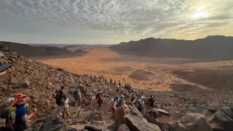 Adam Van de Velde A vast expanse of rocky and sandy desert and a large group of runners following one another in a line. Large hills in the background and a mix of clouds and sunshine in the sky.