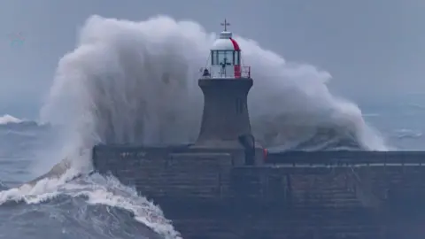 A white, foamy wave rises above the height of the brick lighthouse at the end of the pier. The top of the lighthouse is painted white and red, with a window for the light to shine through.