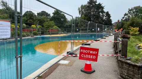 Temporary wire fencing around the edge of a large public paddling pool, which has a blue and yellow bottom. The footpath around the edge of the pool is blocked with red tape, a traffic cone, and a sign reading "footway closed".