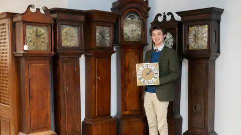 Smartly dressed young man holding a clock face and standing in front of grandfather clocks