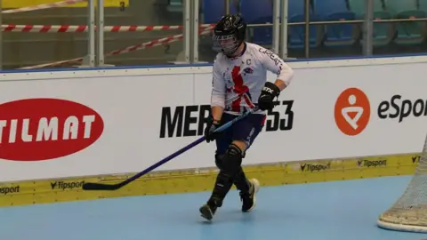 JACOB MANSELL A ball hockey player moves behind a goal on an indoor blue surface. He holds a stick and wears a helmet, gloves and shin pads.