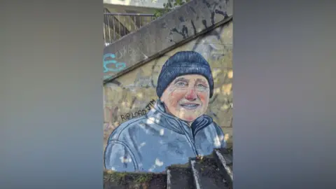 Andy Dice Davies A mural of an elderly man from the chest up, wearing a blue beanie hat and a blue fleece. He is smiling and has a pink nose. Round his shoulders are the words 'RIP Lord Jim'