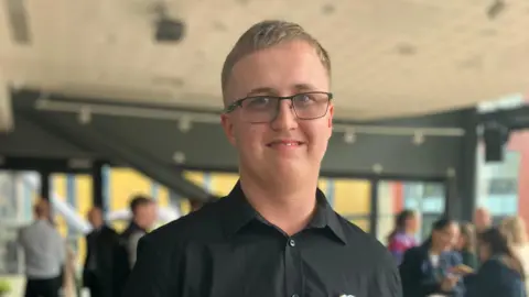 Nathan Fowles in a black shit and glasses smiles for the camera in the presentation room.