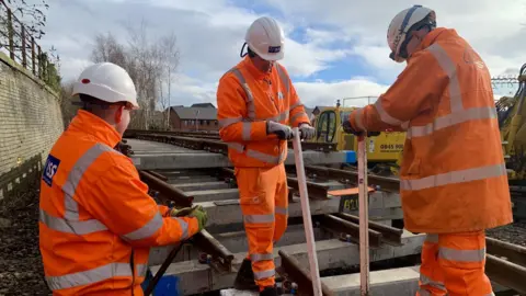Three men in hi-vis work gear and white hardhats work on a piece of railway track in a depot