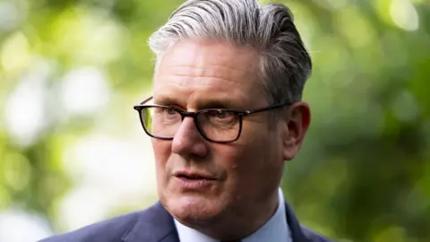 PA Media A man with grey hair and glasses in a close-up shot. He is wearing a dark suit and light-coloured shirt and is visible from the shoulders up. There are trees, out of focus, in the background