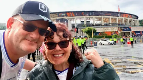 Sarah Lewis A man with an Aston Villa football cap is wearing a football shirt and sunglasses. He is standing next to a woman with red curly hair holding up her fist and smiling to the camera. They are standing in front of a stadium with men in high-vis jackets nearby.