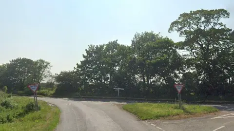 A rural junction, with trees lining one side of the road. A triangular grass verge and two 'give way' signs can also be seen.