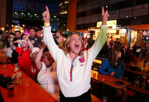 A woman in an England top raises her arms and yells in a packed bar.