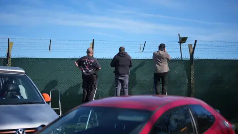 Getty Images Three men stood near a green stand on ladders and peak over barriers. There is a silver and red car at the front of the image and blue sky in the background.