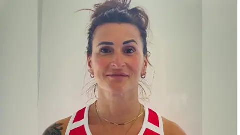 Sabina Falcone-Healey. She has dark hair, which is tied up in a bun. She is wearing a red and white vest. She is pictured against a plain background and is looking directly at the camera and smiling.