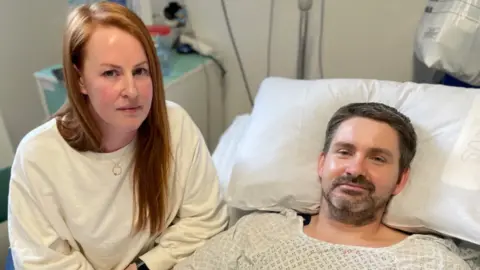 A man with short brown hair and facial hair, wearing hospital gown and lying in a hospital bed. Beside him is a woman with long red hair, wearing a white jumper. They are both looking at the camera with serious expressions.