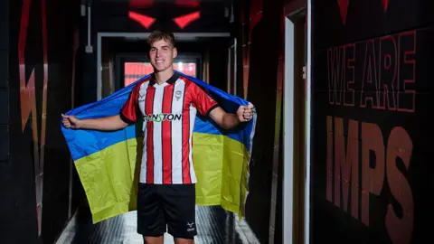 A man standing in the tunnel at a football stadium. He is wearing Lincoln City's red and white colours, and has a yellow and blue Ukrainian flag around his shoulders. He is smiling.