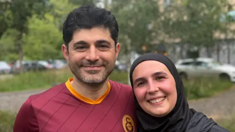 Elliot Deady/BBC A man and a woman smiling straight at the camera. There are trees and parked cars behind them. The man is wearing a burgundy and orange T-shirt and the woman is wearing a black hijab.