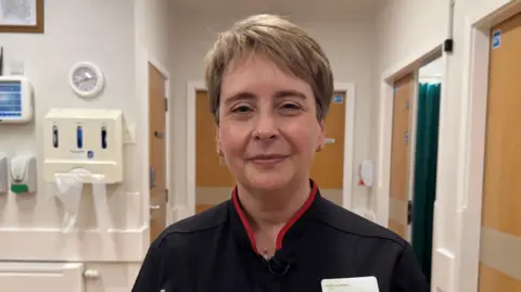 Gill Findley is standing on the hospital ward, looking directly at the camera and smiling slightly. She has short blonde hair and is wearing a dark blue and red uniform. There is hospital equipment behind her including hand sanitiser dispensers.