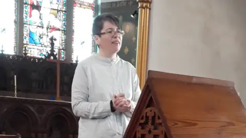 Jen Bressers Jen Bressers - a short-haired woman with glasses, gives a sermon from the pulpit of a church - an altar and stained glass window behind her. She wears a light grey shirt.