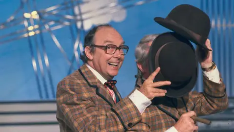 Getty Images Eric Morecambe is grinning as he holds his hat to block Ernie Wise's face during a dance routine