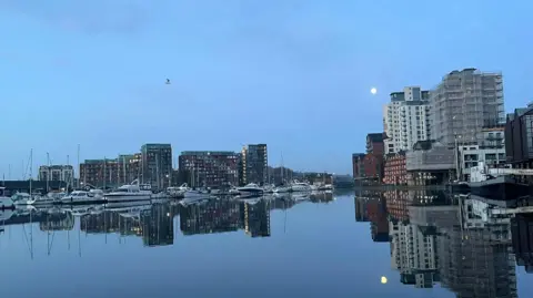 Connor Bennett/BBC A marina showing blue grey water under a blue grey sky. It is ringed with high rise flats and on the left side, power boats are moored up. The boats and flats are reflected in the mirror-flat water. 