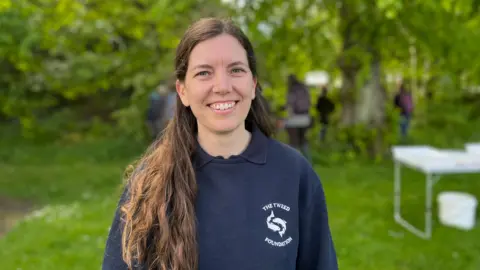 A woman with long brown hair, wearing a dark blue top, smiles at the camera
