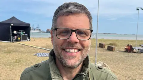 Richard Daniel/BBC Amit Lahav stands in a grassy area used for the performance. He Is wearing glasses and a khaki green jacket, and he has grey hair and a grey beard. He is smiling at the camera