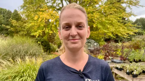 Kristyna Kay-Clough, smiling for the camera. A garden with several plants and a large tree with yellowing leaves are visible behind her.