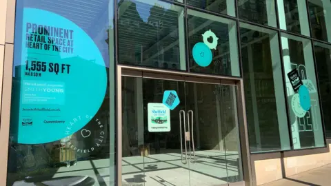 A large glass fronted unit in a city centre is empty inside. There are double doors and windows which have turquoise stickers offering the unit for rent