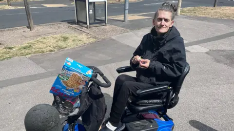 BBC A woman with dark, greying hair, pulled back into a hair band, sits on a blue mobility scooter with shopping in the front basket. She wears dark clothing. 
