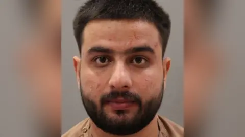 Police custody image of Sultani Bakatash. He has black hair and beard and is wearing a beige t-shirt.