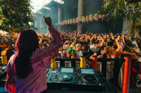 A woman wearing a pink jacket DJs at an Alfresco Disco event in Bristol with a packed crowd dancing in the background