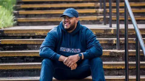 A man wearing a blue baseball cap and blue tracksuit is sitting on some steps outdoors