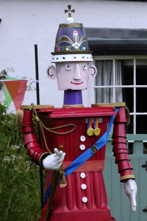 Julie Sanders A model of a man made from plant pots in a red uniform, medals and a cross on his hat. There is a building behind it with a window, a gate and bush.