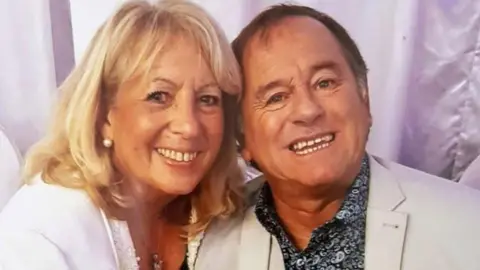 Family photograph Lynn Trevallion with shoulder length blond hair wearing a white jacket with the late comedian Duncan Norvelle who has short grey hair and wearing a cream jacket and black and white patterned shirt.