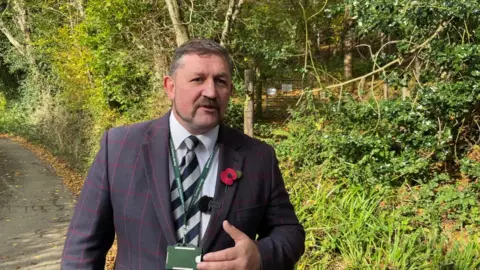 Councillor Dermot MceGeough stands, facing the camera and wearing a pinstripe blue and red jacket, blue and white striped tie and white shirt, with a poppy on his jacket lapel. He has a moustache and wears a council lanyard. Behind him is a single track road and shrubbery. 