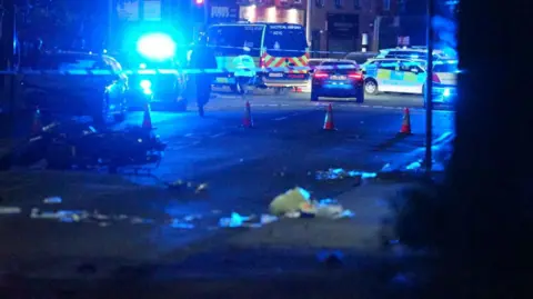 Night time shot of police cordon with multiple police vehicles close by. A motorbike can be seen vaguely in the left hand side of this picture.