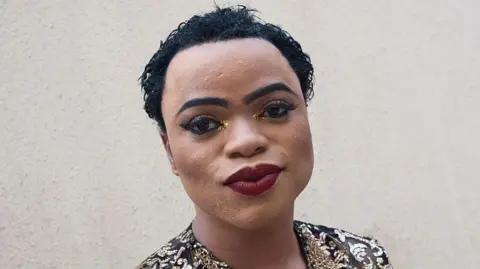 AFP A close-up shot of Bobrisky wearing red lipstick, glitter eyeshadow and a gold and black top. 