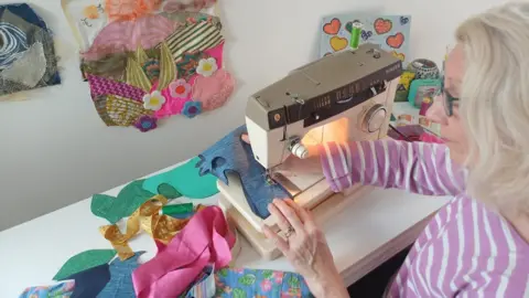 The Wilson Jennifer sews colourful pieces of fabric together at a sewing machine. Some small works of hers are displayed on the wall behind her. She wears a pink and white striped top and blue-framed glasses. 