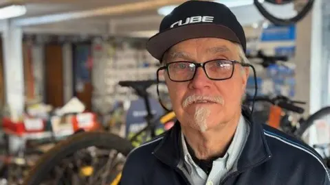 Daan van Belzen of Bikeseven, of Longtown. He has a white goatee, blue eyes and is wearing glasses with a dark rim. He is wearing a black baseball cap with white lettering and a blue top with white and light blue stripes on the shoulder-line. He is in the bike shop and the background is blurred.