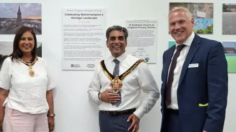 Martin Phillips/Historic England Three people stand in front of photographic exhibits and captions on a white wall in an exhibition space, smiling at the camera. A woman with long dark hair is on the left wearing a white top and small mayoral chain. The man in the middle has a white shirt, grey tie and larger chain, and the man on the right is in a blue suit, white shirt and dark tie.