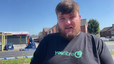 Jonny Humphries/BBC Eddie Puskepalis, in his mid-20s with brown hair and a brown beard and wearing black t-shirt with a 'Youfibre' broadband logo, speaks to the camera with police tape and blue tarpaulin sheets next to a bus-stop behind him.