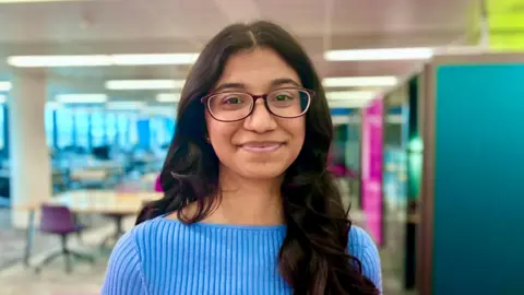 Bhuvana Chilukuri with long dark brown wavy hair and glasses wearing a pale blue ribbed top and standing with a university library environment behind her